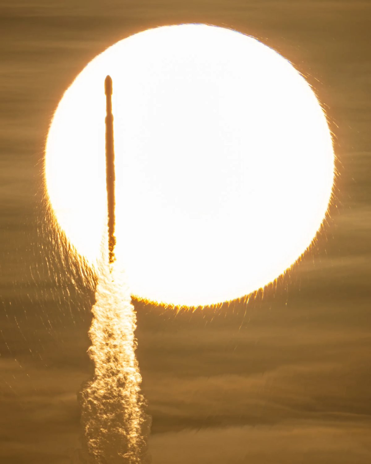 Silueta del cohete Falcon Heavy subiendo frente al disco del Sol, con su estela iluminada.